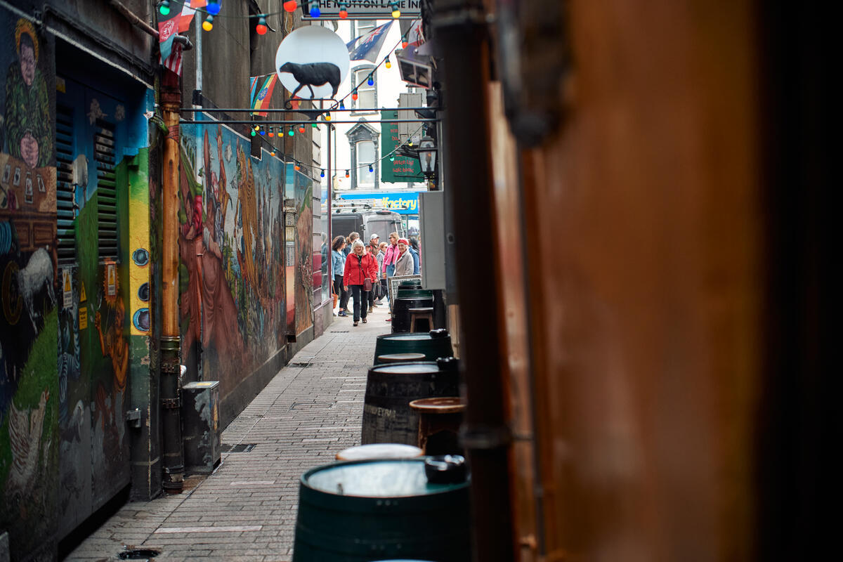 The narrow entrance to Mutton Lane in Cork city, with colourful murals, hanging lights, beer barrels and people walking through the laneway beyond.